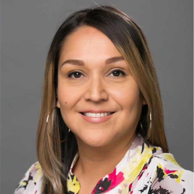 Apache Danforth smiling and facing the camera. She wears hoop earrings and a floral shirt.